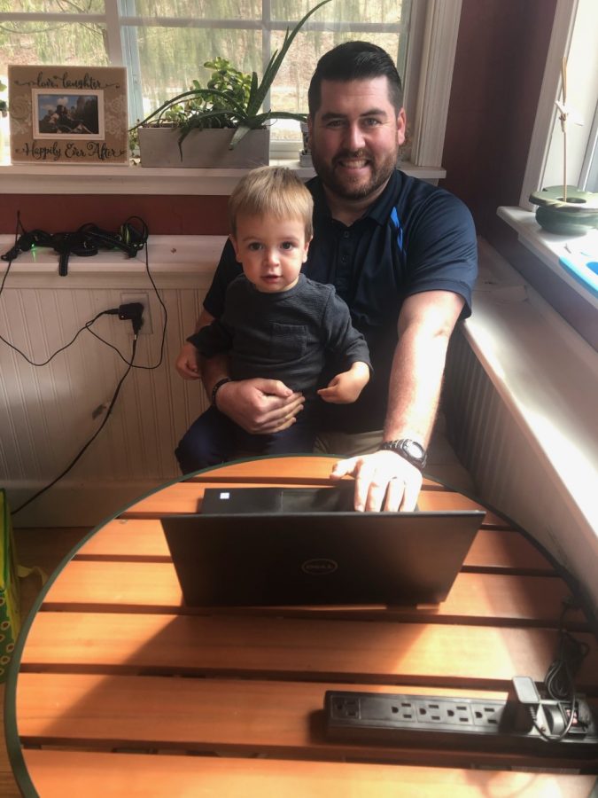 Mr. Zelger holding his 2-year-old son, Nathaniel, also typing on the laptop. It is seen that he is juggling, by having a child and doing Zoom classes.