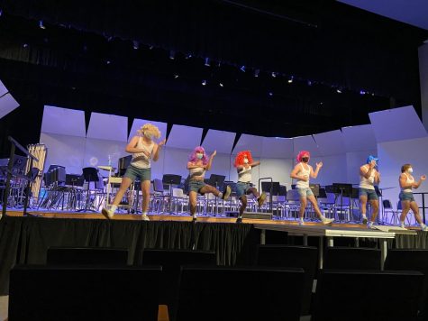 6 contestants, dressed in white tanks tops, jean shorts, and colorful wigs, dancing on stage as a group.