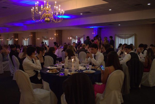 Students sat around a blue circular table with white chairs, eating dinner in their Prom entire in a room dimly lit by a golden chandelier and blue lights.