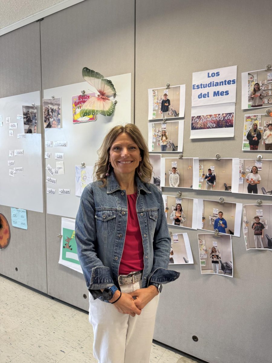 A middle aged women in a red shirt and jean jacket with white jeans standing in a classroom with pictures of her students on the walls.
