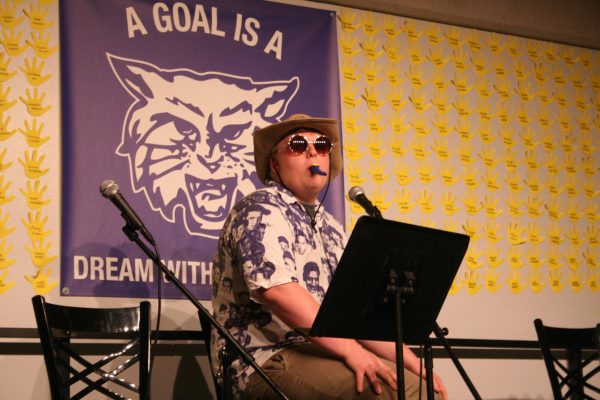 A boy on stage dressed in a blue and white button up shirt, hat, and circular glasses, placing the kazoo without his hands.