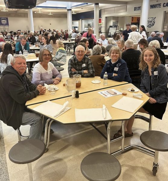 4 senior citizens sitting down with a student council member at a table, connecting with one and other while eating a snack and drinking a beverage.
