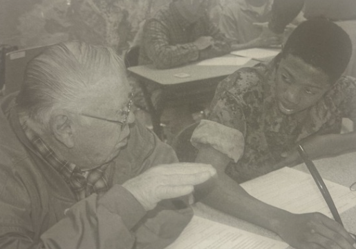 An elderly man and young male student, dressed in an JROTC military uniform, sitting down talking while the young male student is completing an assignment.