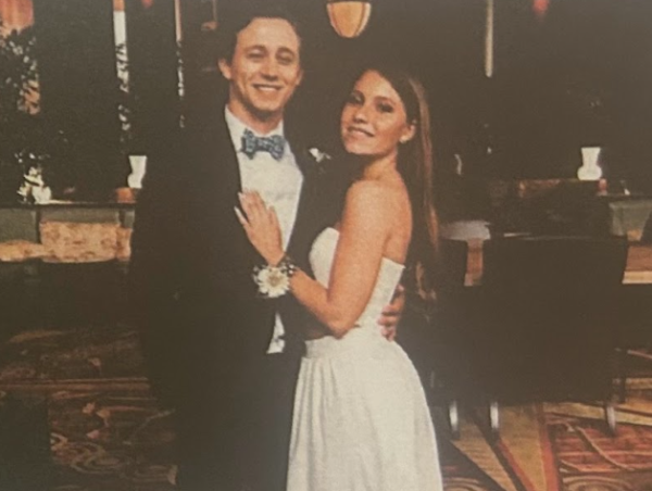 A boy, dressed in a black tuxedo with a blue bow tie, standing next to a girl dressed in a 2-piece white gown, smiling.