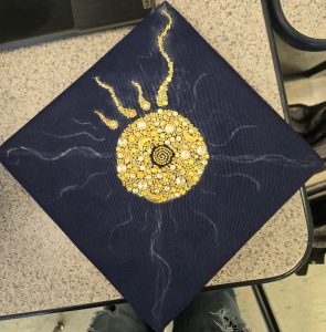 Navy blue graduation cap decorated with white and yellow gems with a sun design outlined in white pencil.