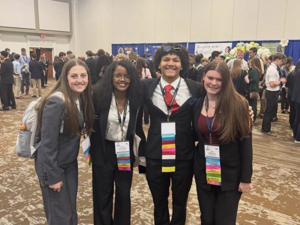 Four high school students pose with hands behind each other’s back in a line and smile for a picture.