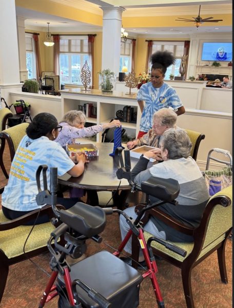 Three sitting senior citizens and two high school students, one sitting one standing, gather around a round table to play a table board game.