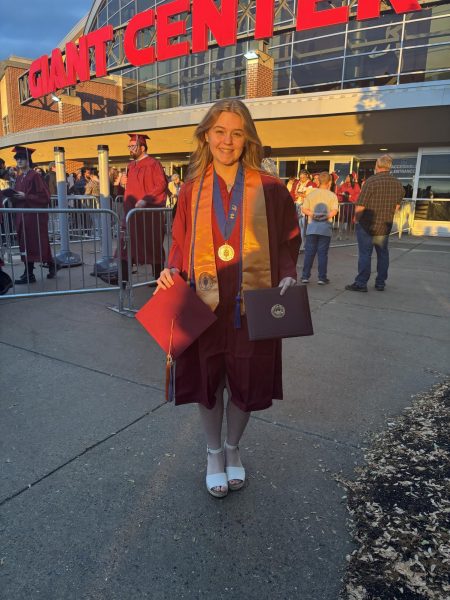 A girl wearing a red graduation gown holding her cap and degree.