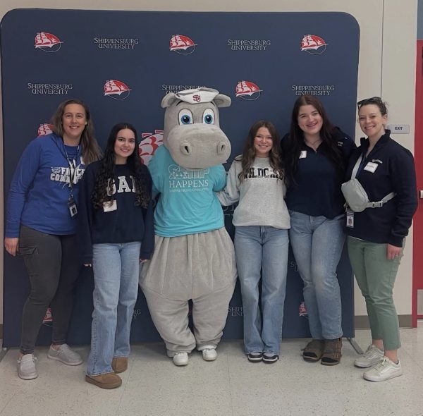 Students, teachers, and a hippo mascot with their arms rapped around each other standing in front of a blue background.