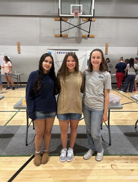 Three girls with their arms around each other standing in front of the a Dallastown Educators Rising board inside of a gym.