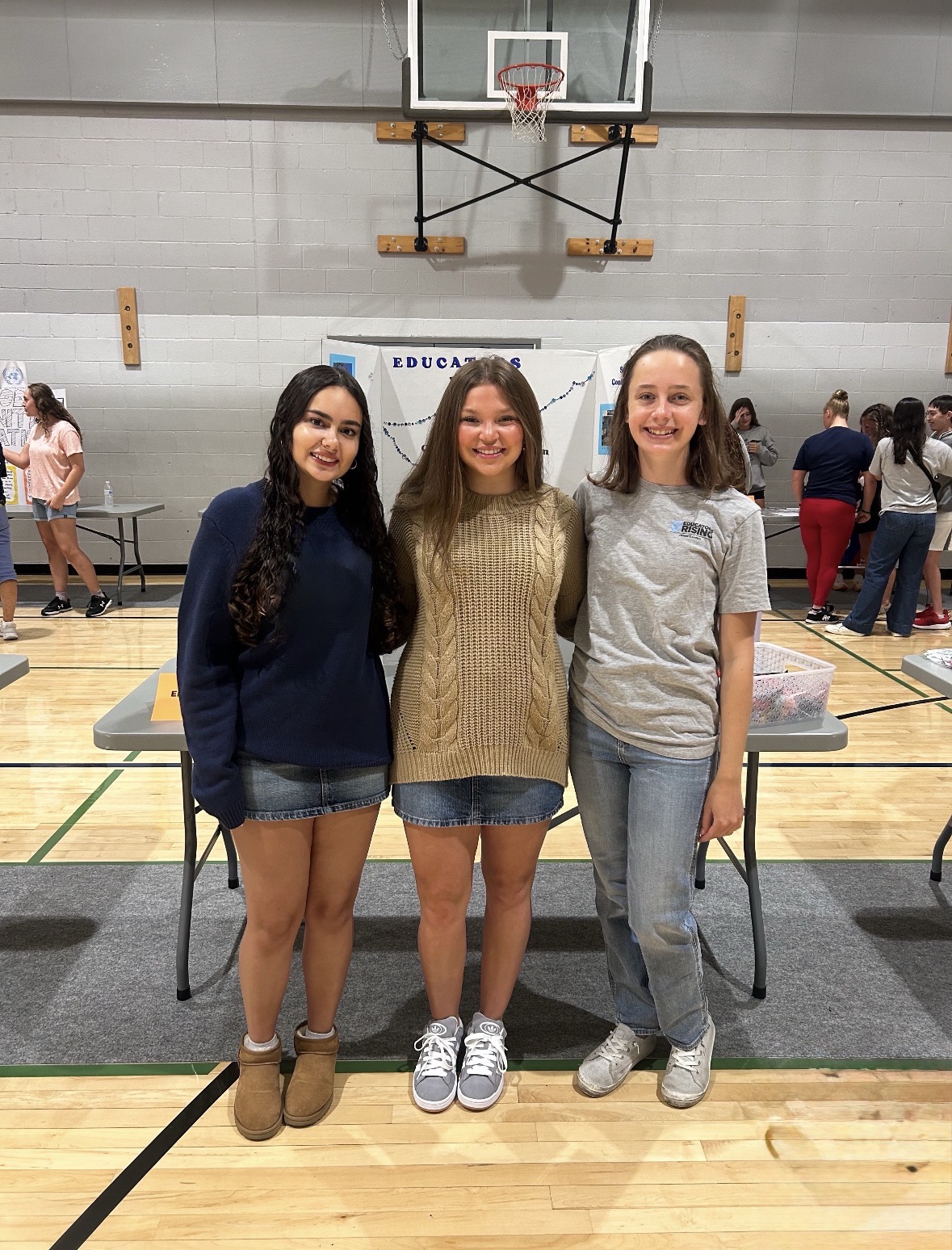 Three girls with their arms around each other standing in front of the a Dallastown Educators Rising board inside of a gym.