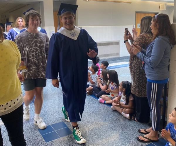 A line of Dallastown graduates, dresses in a navy-blue cap and gown, walk down a school hallway lined with teachers and younger students.