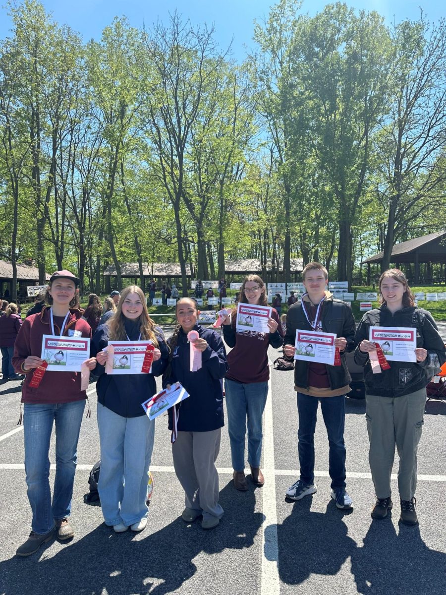 Six high school studnets wearing red envirothoin shirts and assorted jackets standing in a line with medals around their necks, holding paper certificates and pink ribbons.