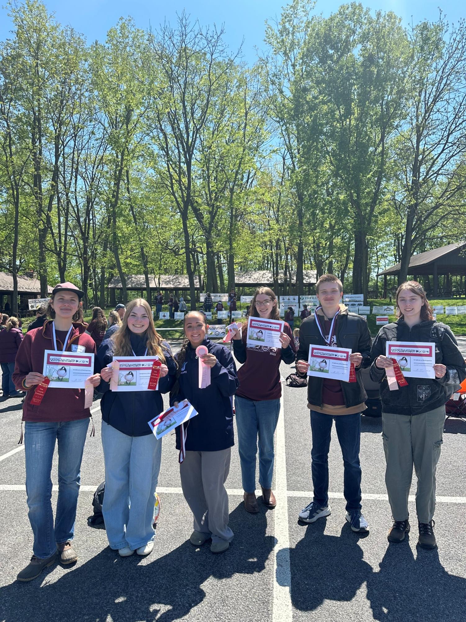 Six high school studnets wearing red envirothoin shirts and assorted jackets standing in a line with medals around their necks, holding paper certificates and pink ribbons.