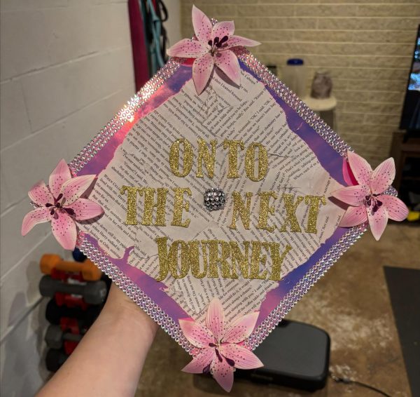 Graduation cap decorated with pink flowers on each corner, pink holographic and gems along the side, torn book pages in the middle with "onto the next journey" spelt in big gold letters.